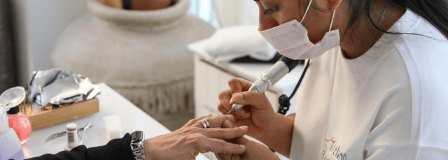 Nail technician wearing mask applying nail treatment with precision tool to client hand in professional salon setting