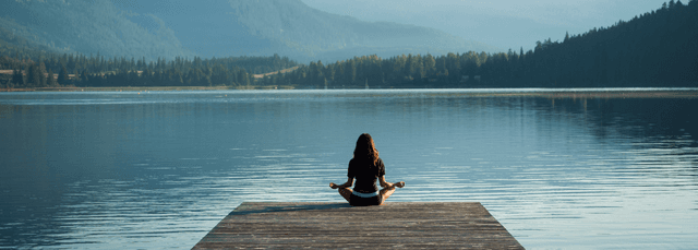 Woman meditating in lotus position on wooden dock overlooking serene mountain lake surrounded by forest