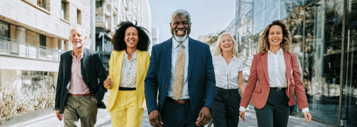 Diverse team of five business professionals in suits walking confidently together outside modern office building