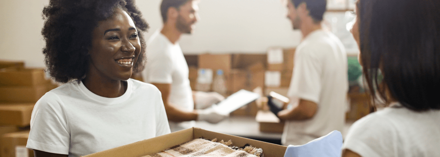 Smiling volunteer in white shirt holding donation box with team of volunteers sorting items in background